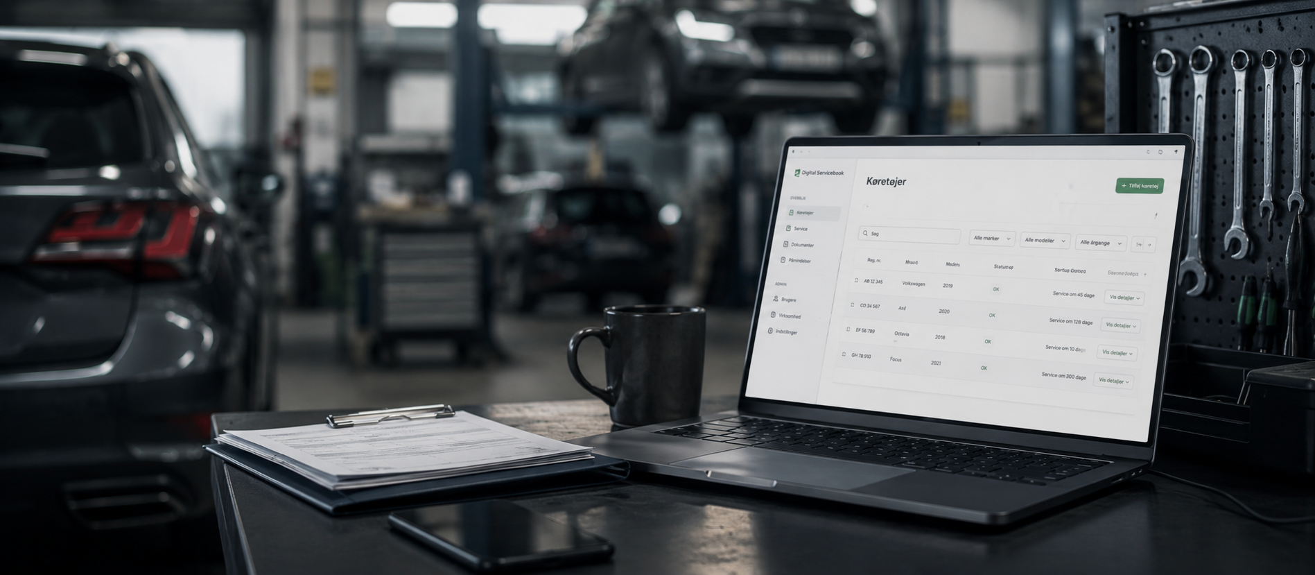 A laptop showing the Digital Servicebook app on a workshop bench, with cars on lifts in the background